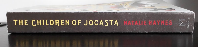 A photo of the spine of a proof copy of The Children of Jocasta. It is black, with the title in yellow and the author's name in red. The book lies on a black desk.